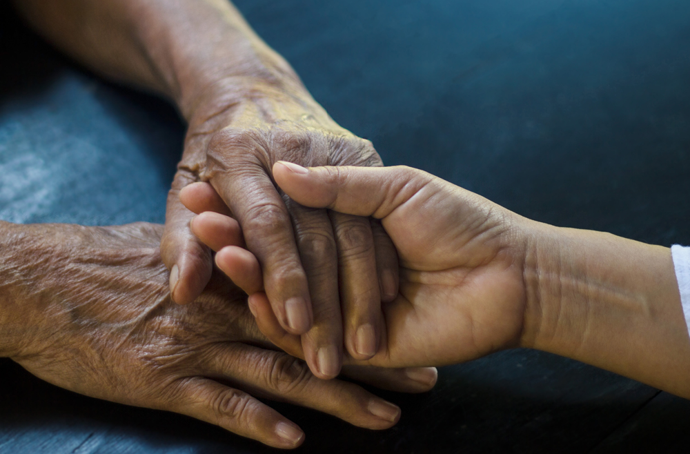 A close-up photo of holding hands in which a hand from a younger person holds one of the pair of hands of an older person.